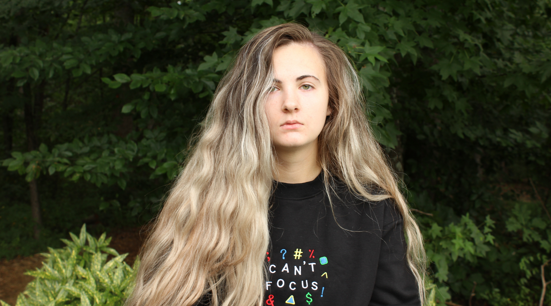 Woman with long blonde hair wearing a black shirt with colorful text in front of green foliage